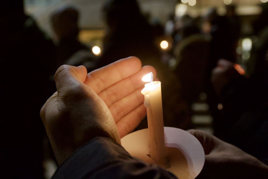 A hand holds a lit candle while others also holding candles are seen, blurred, in the background