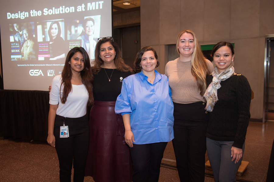 Photo of five women posing in front of a wall-mounted video screen, with a lecture slide that says "Design the Solution at MIT"