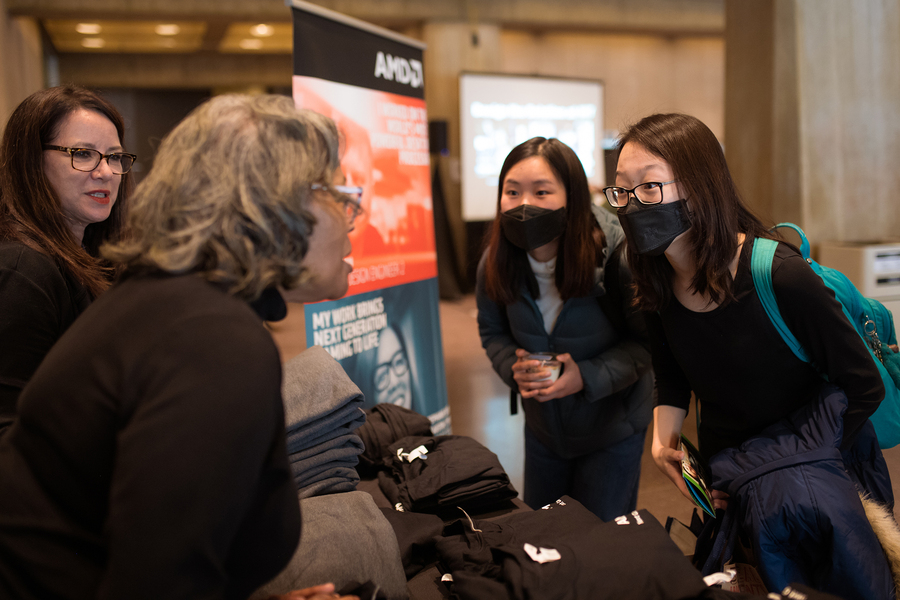 Photo of two women, leaning forward to speak with two representatives staffing a table with piles of T-shirts
