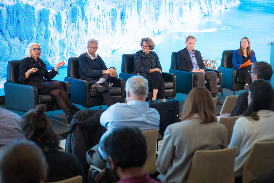 Five panelists sitting in armchairs side by side on a stage, facing rows of audience members