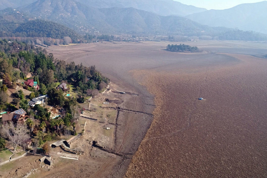 Aerial view of a brown lake with large banks of dried mud. There is a single boat on the water, and a few houses surrounded by trees on a patch of land.