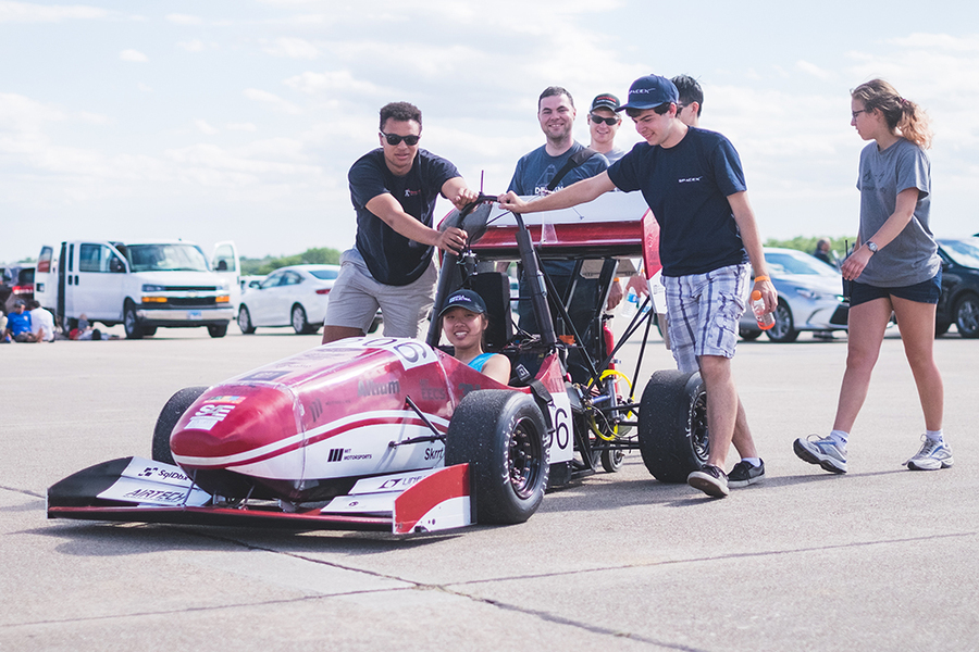Photo of Cheyenne Hua in motorsports vehicle with several teammates standing behind her