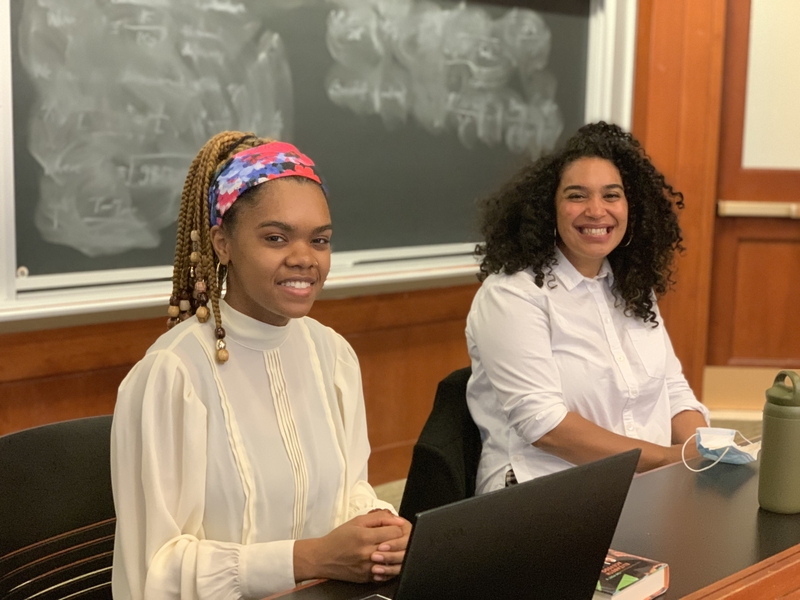 Photo of two women seated before a blackboard, one with a laptop