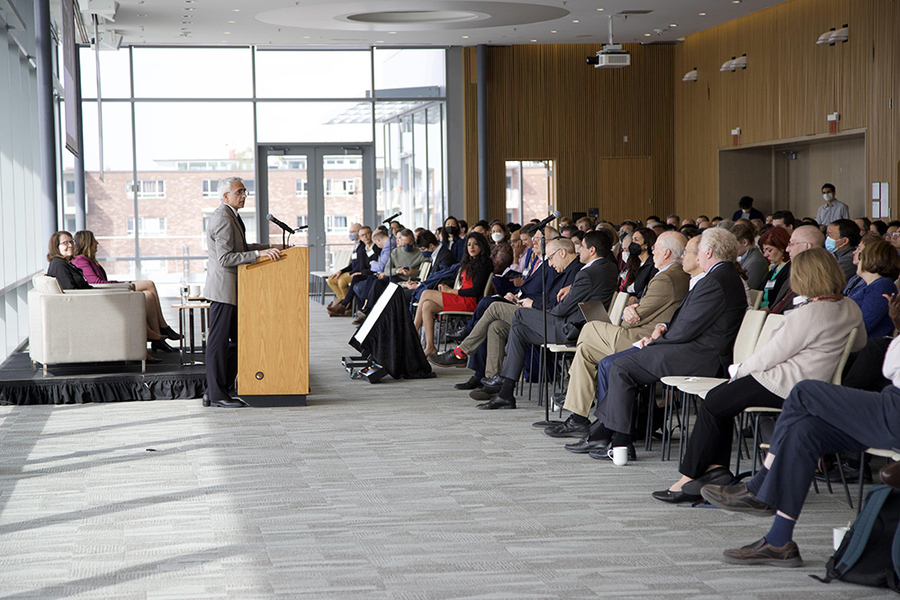 Photo of a man speaking at a podium in front of an audience in a room with floor to ceiling windows
