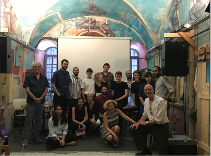 Photo of Mark Jarzombek posing with 16 students in a room with a colorfully painted arched ceiling. They are all posing in front of a pull-down media display screen.