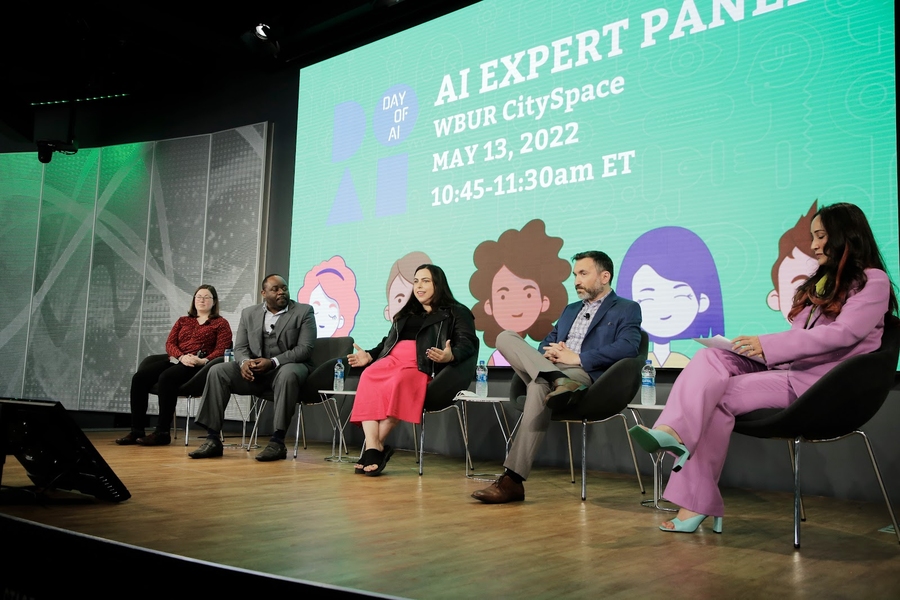 Photo of four panelists and one moderator onstage. A screen behind them says "AI Expert Panel" with five cartoon images of young people's faces