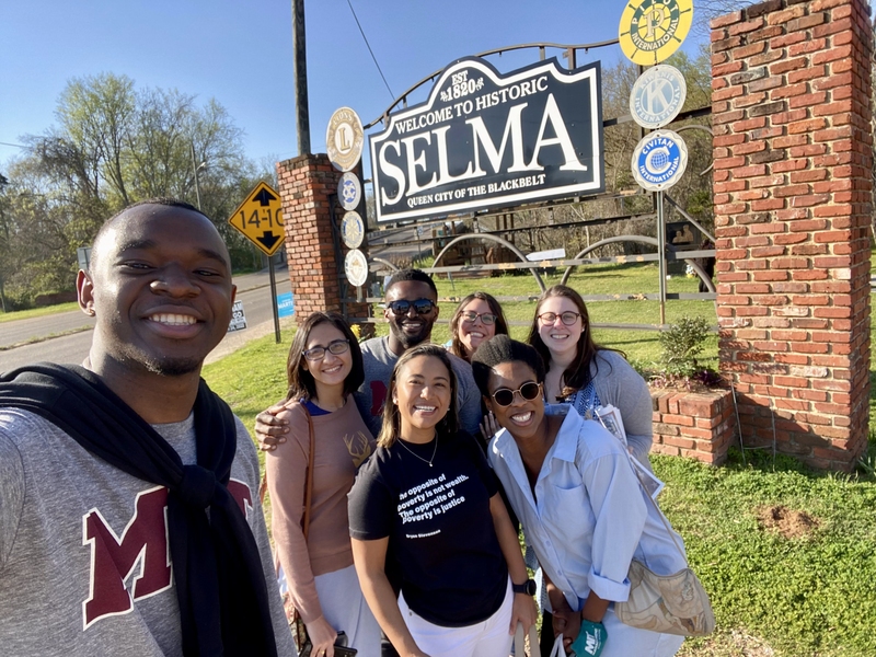 Photo of seven students posing in front of a large road sign that says "Welcome to historic Selma, queen city of the blackbelt"