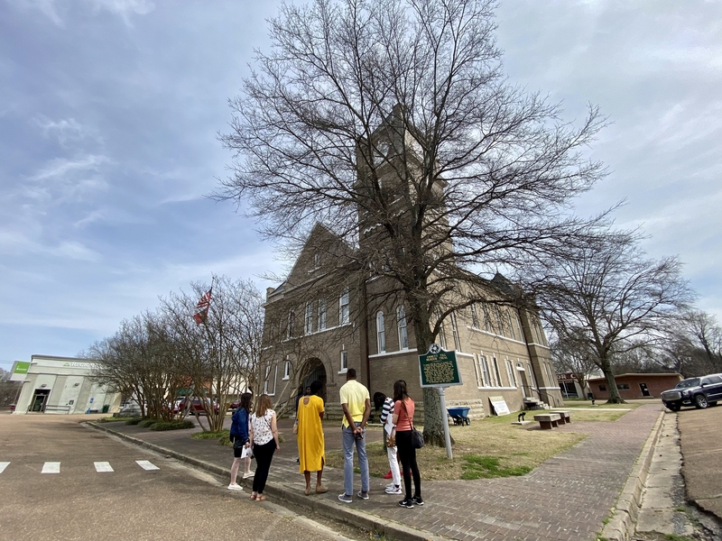 Wide-angle photo of students in standing outside in front of a courthouse. A permanent sign posted outside reads "Emmett Till Murder Trial"