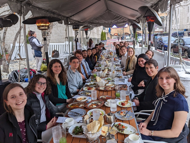 Approximately 25 students sit on either side of a long dining table that is outdoors and covered by a tent