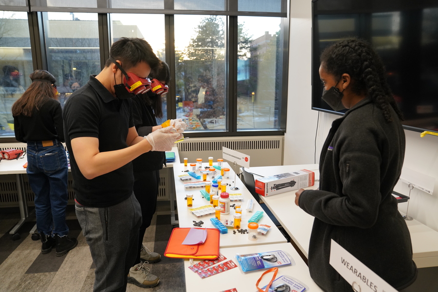 Students wear red limited-vision goggles while trying to identify medicines. There are more than two dozen medicine bottles and containers on a table in front of them.