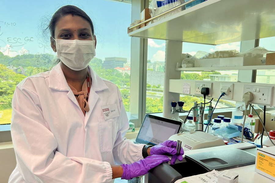 Photo of Shruthi Pandi Chelvam in lab, using a piece of lab equipment in front of a large window with treetops and some buildings in the background