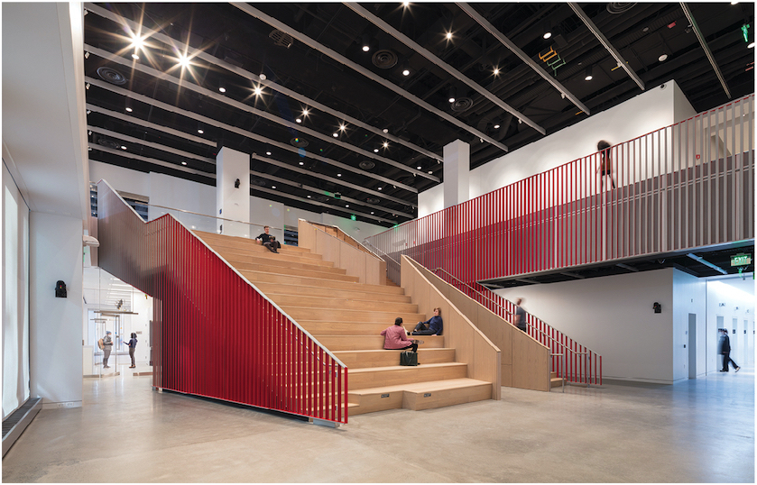 Interior photo of a two-story auditorium, which is a large, wide wooden staircase with red railings on the side. Three people are seated while others walk around the adjoining spaces.