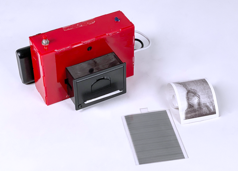 Photo of KineCAM, a bright red plastic box with smaller black plastic attachments. Also seen are a sheet of acetate with gray lines and a sheet with the outline of a woman.