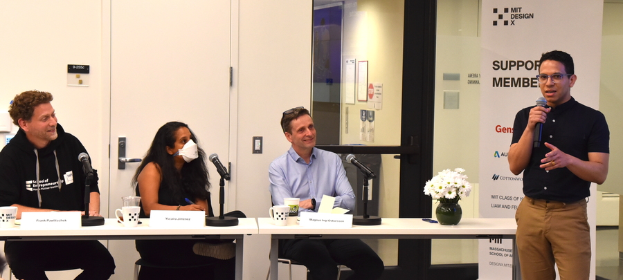 Photo of three people seated at a table listening to a fourth person, standing and speaking into a microphone
