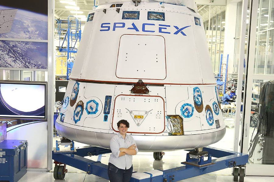 Photo of Joy Dunn standing with her arms crossed, smiling, in front of Dragon spacecraft at SpaceX.