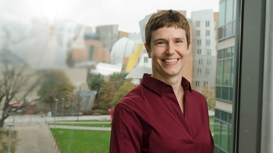 Amy Keating stands in a red button-down blazer in front of her office window at MIT.