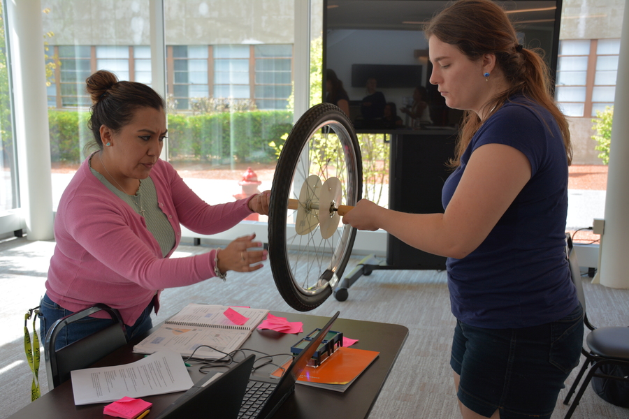 Two teachers participating in the LLRISE program hold a bike wheel between them, turning it and measuring its movement with a small radar on a table.