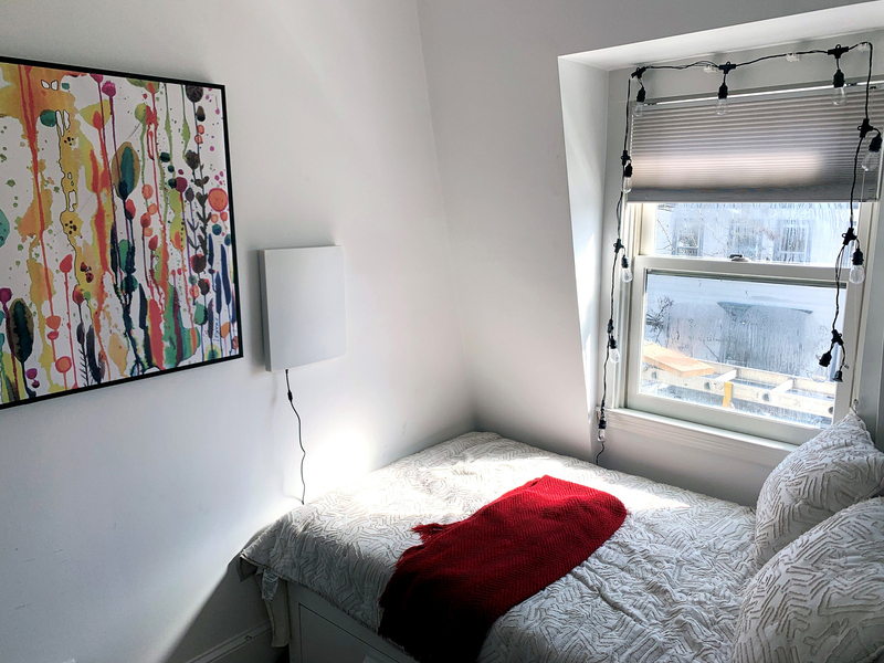 Bedroom photo showing a bed, a painting, a window, and a white box hanging next to the painting.