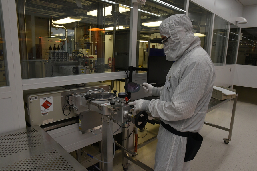 A researcher wearing a white, protective lab suit inserting a silicon wafer into a large stainless steel apparatus in the MIT.nano cleanroom.
