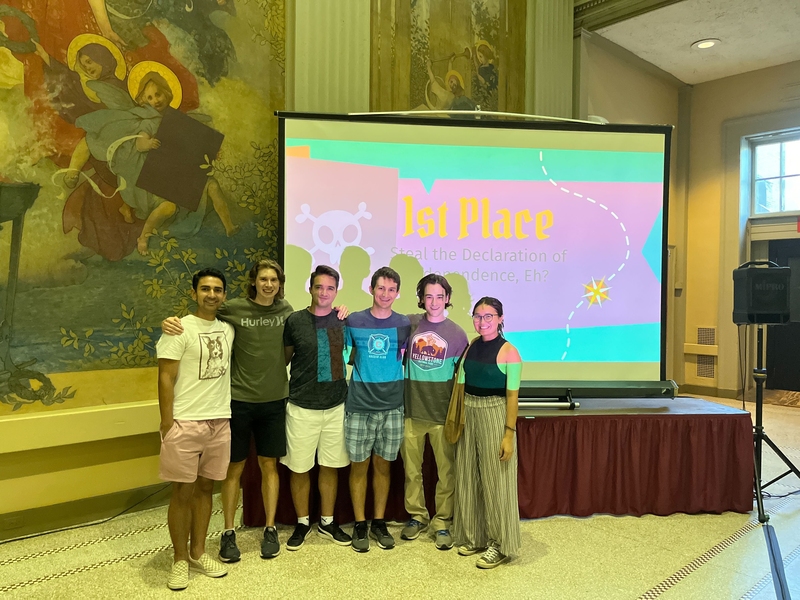 Six graduate students pose together in front of the projection screen that reads, “1st Place” and their team name, “Steal the Declaration of Independence, Eh?” The screen is set up in front of wall-length murals in MIT’s Morss Hall.
