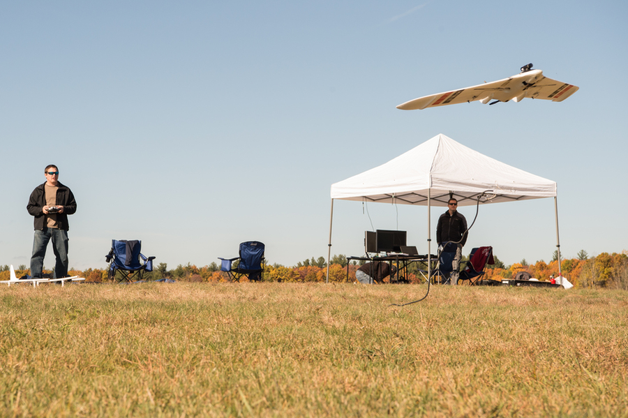 a researcher flies a small drone via remote control. He is standing in field; a tent is set up behind him, where more researchers are working.