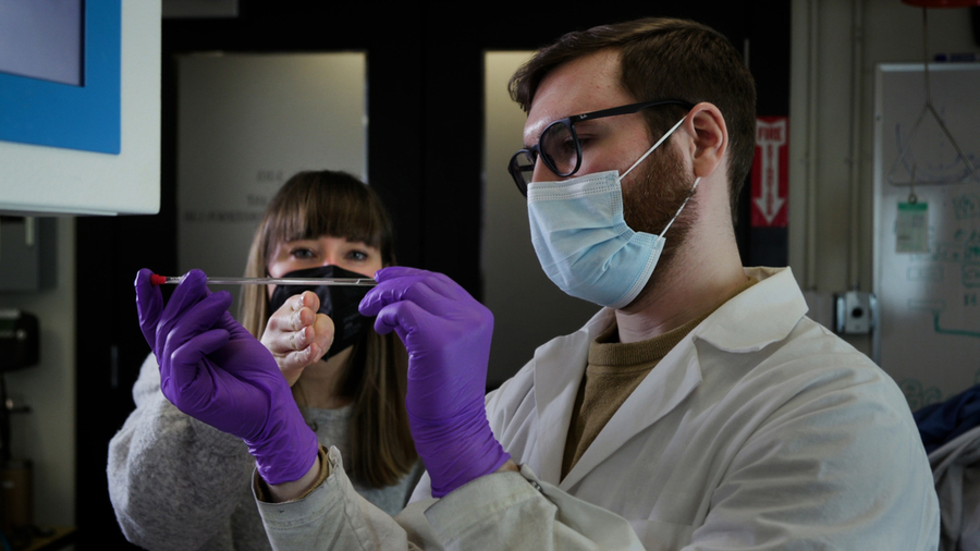 Photo of a man in lab gear examining a pipette full of liquid under a woman’s guidance