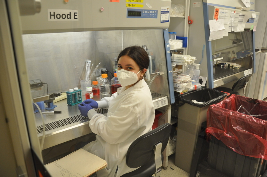 Alejandra Rosario sits at a lab bench wearing a white coat and purple gloves.