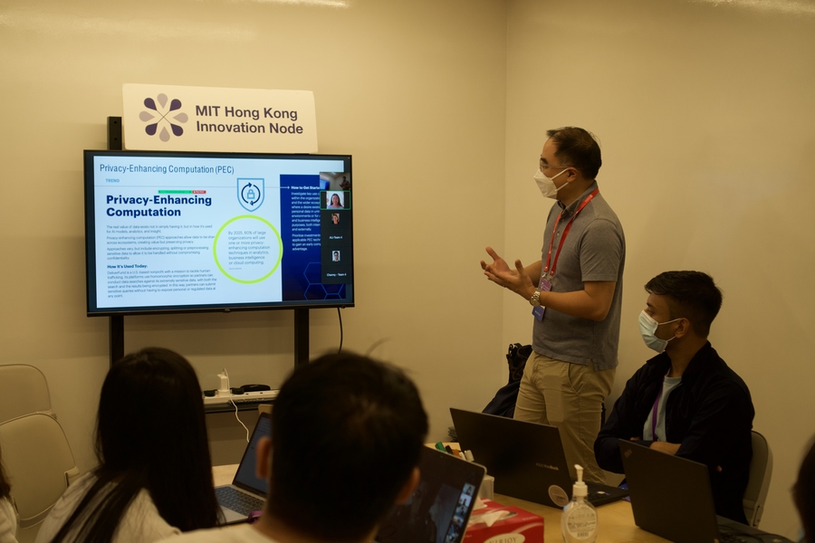 A man stands next to a table with several people; they are all watching a presentation on a large screen on the wall that is labeled with "Hong Kong Innovation Node." The screen itself mostly unreadable text with the header "Privacy-Enhancing Computation (PEC)." Images representing remote participants appear at the right of the screen.