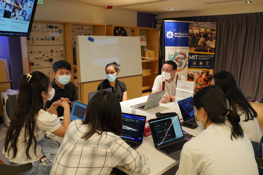 In a small room, a team of students sits around a table, in conversation. They are all wearing face masks and most have laptops open.