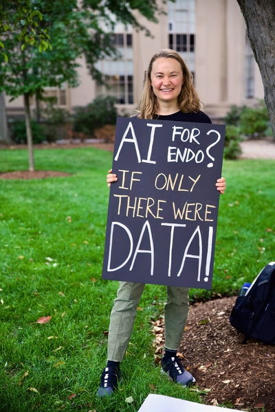Kira Buttrey stands in a grassy area, holding a painted sign reading "A.I. for endo? If only there were data!"