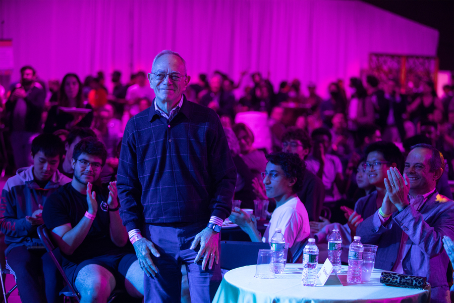 In a large room with at least 100 people in view, MIT President L. Rafael Reif stands as others seated at tables next to him clap. Some in the background are standing.