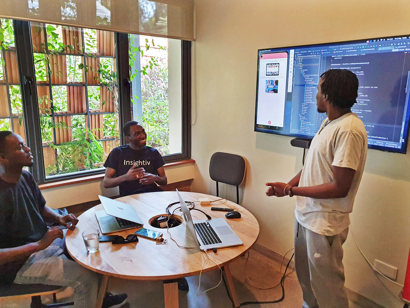 Two men sit at table while third man presents a slide on a digital screen
