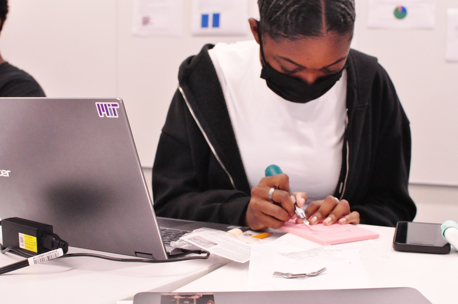 Photo of a teenage girl wearing a mask and creating a rubber block stamp. A laptop is propped open beside her with an MIT sticker on it.