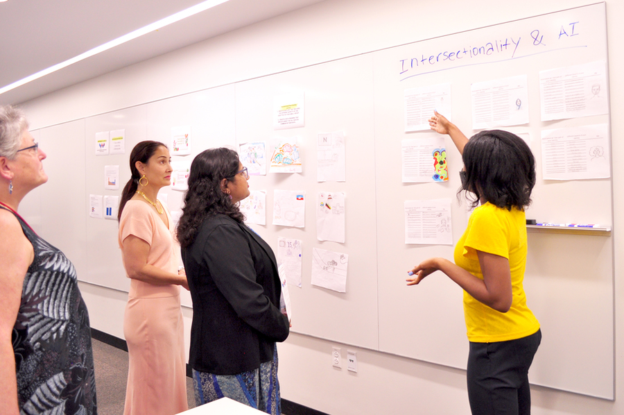 Photo of Raechel Walker gesturing to a whiteboard with "Intersectionality & AI" written on it and student projects taped underneath. Sumbul Siddiqui, Cynthia Breazeal, and another woman from the City of Cambridge Office look on.
