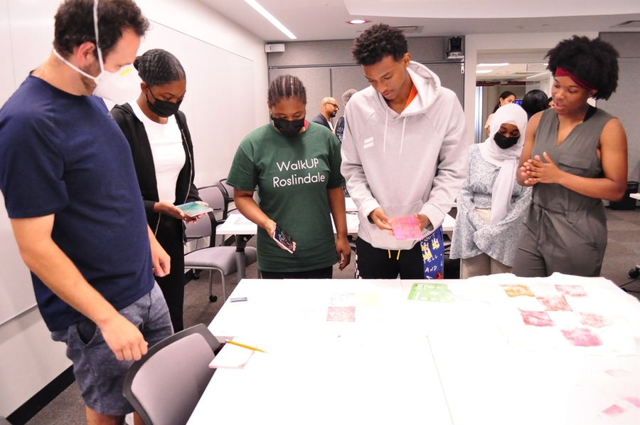 4 students and 2 adults look at a T-shirt laid out on a table with multiple stamp designs printed on it. The students are holding block stamps in their hands.