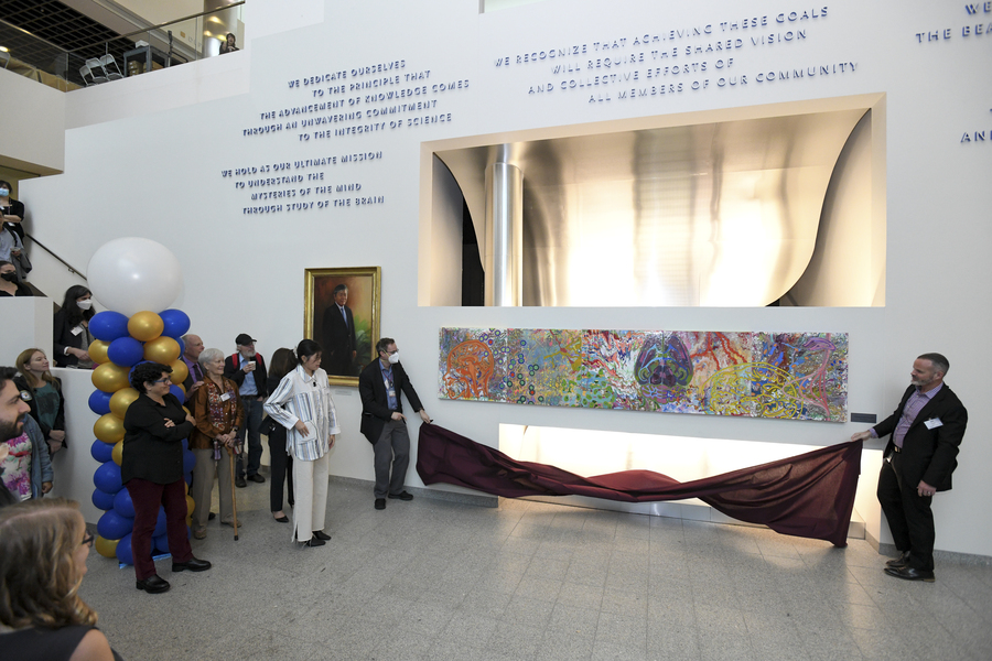 In the lobby of The Picower Institute in MIT's Building 46, two men hold a cloth under a wide, colorful painting composed of neuroscience imagery. Mila Sheng, the artist, stands nearby. To her right is Nergis Mavalvala. Standing behind them are many onlookers and a tall column of blue, gold and white balloons.