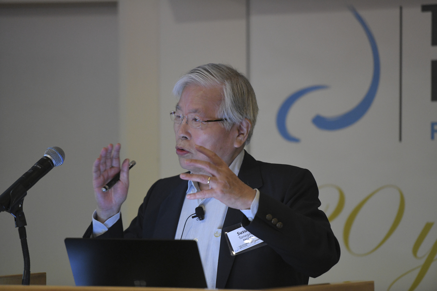 Susumu Tonegawa stands at a podium with the Picower Institute logo behind him. His hands are raised in an emphatic gesture as he makes a point during a talk.