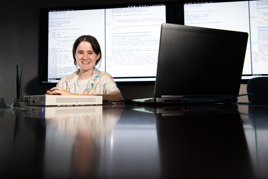 Mya Gordon sits behind a computer and receiver. Large screens with programming text are in the background.