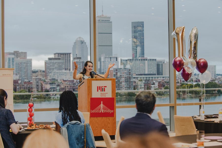 Taylor Patskanick speaking with her hands in the air at podium. The Charles River and Boston skyline are in the background, while balloons float from a table beside her.