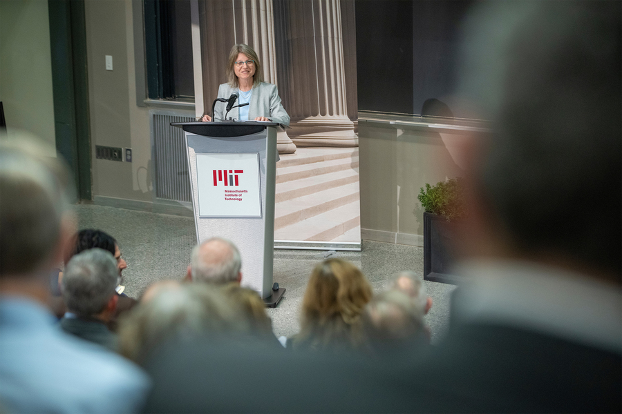 Sally Kornbluth speaks at a podium in front of a blackboard, while audience members watching are seen in the foreground