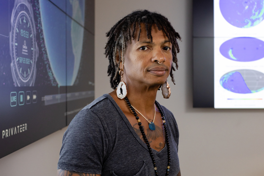 Moriba Jah stands facing the camera against a grey wall. Behind him are two large screens showing images containing maps and a stopwatch