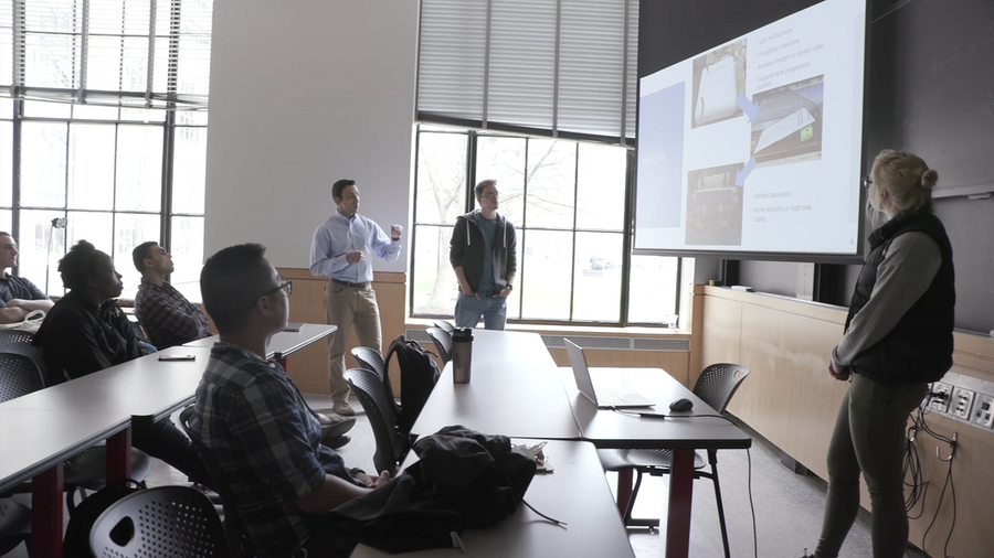 Woman stands in front of screen and black board, presenting a slide to students in a sunlit classroom.