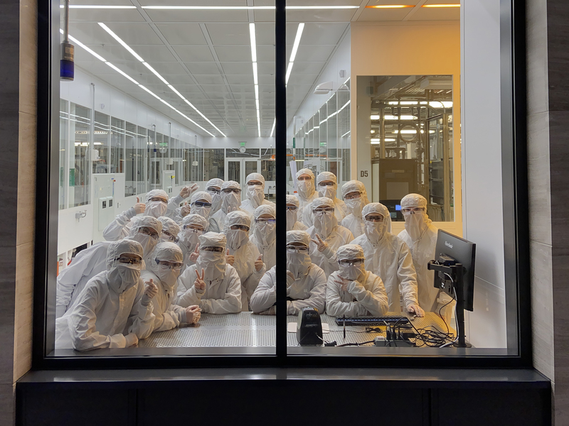 A large group of students, wearing full-body white lab suits, posing in the window of the MIT.nano cleanroom, seen from the outside.