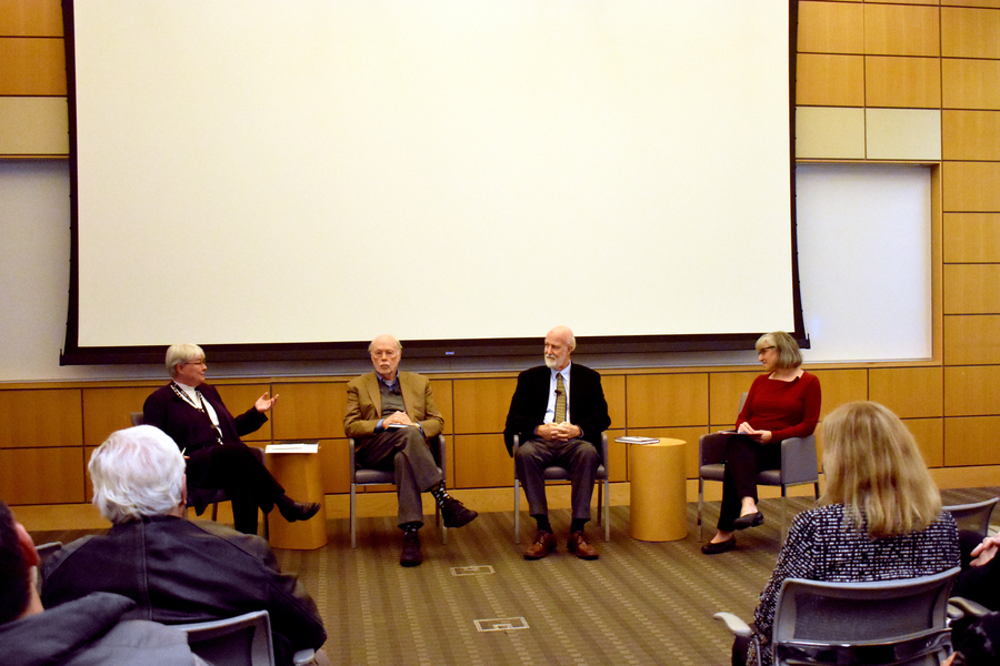 Four people sit facing an audience. There is a large pull-down projector screen behind them.