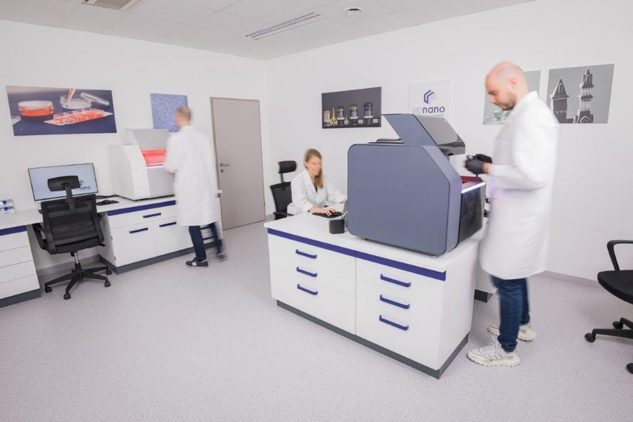 Technicians in lab coats with blurred faces work in a laboratory with an UpNanoNanoOne 1000 printer.