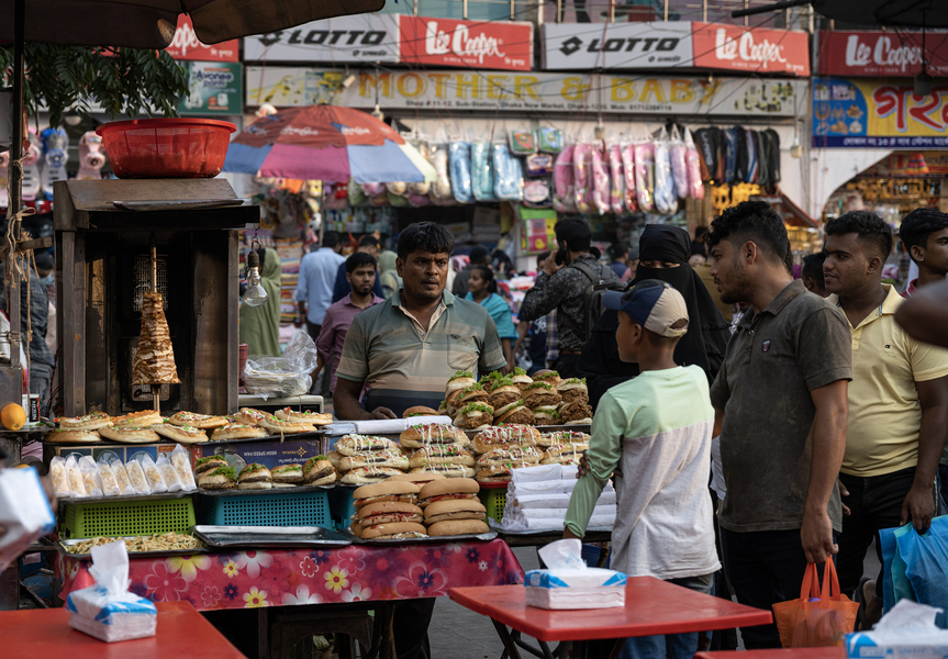 A colorful market stall is shown in a bustling street of Dhaka, Bangladesh. In the foreground, a customer surveys an array of sandwiches; in the background, umbrellas and packages of colorful clothing are visible.