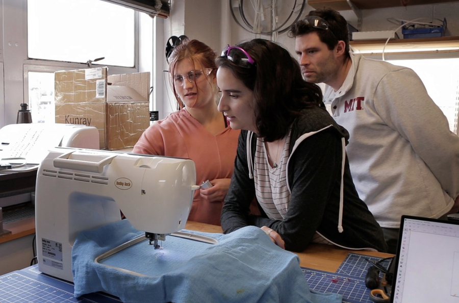 Two women and a man gather around a sewing machine with blue fabric. 