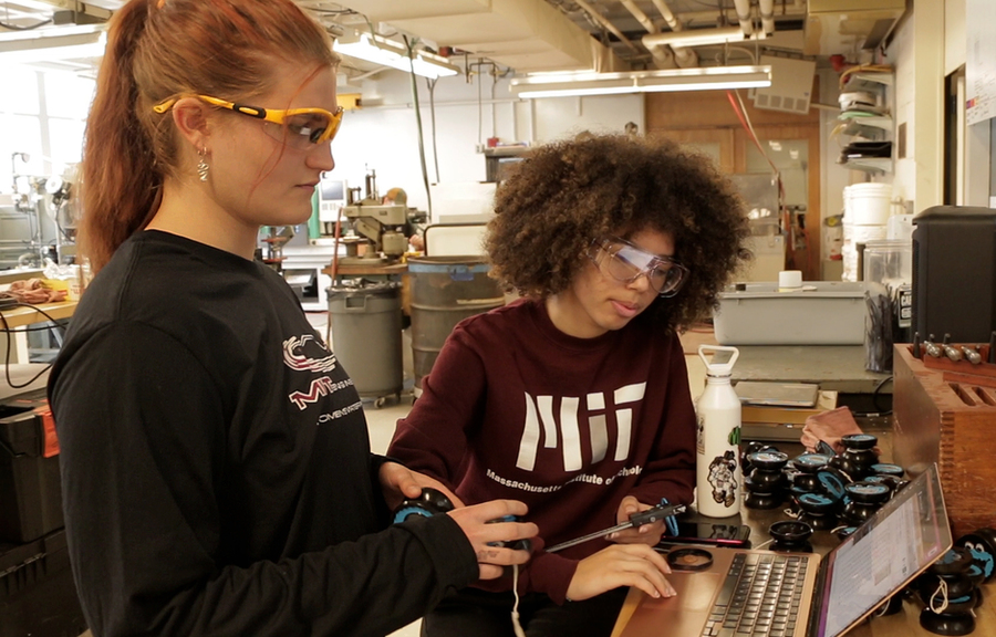 Two women wearing protective googles stand in front of a computer and desk with a pile of yo-yos in the lab. 