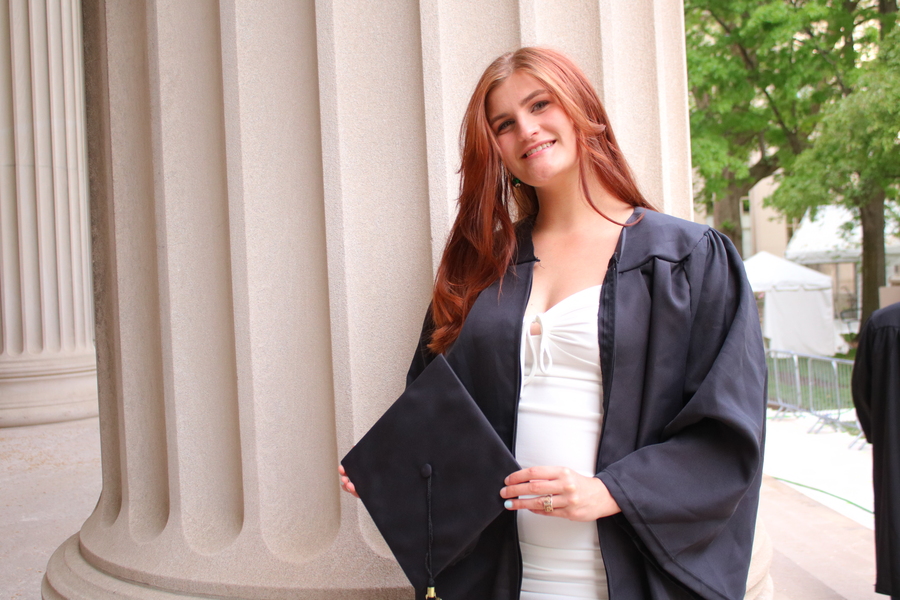 Emily Satterfield in white dress and graduation robes poses in front of large column 