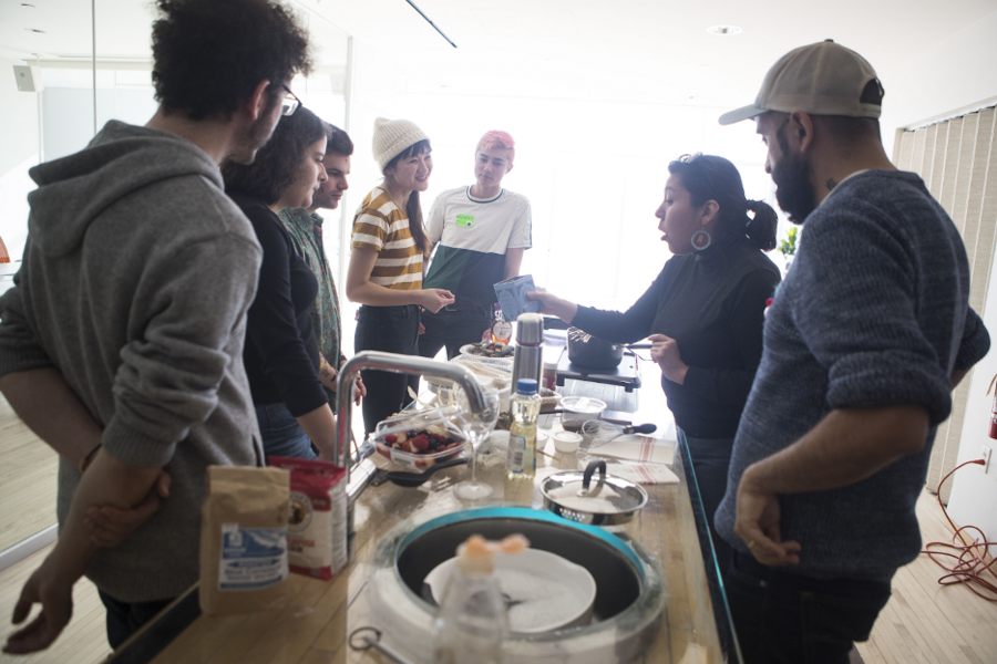 Half a dozen people stand around a food prep counter while a seventh demonstrates something